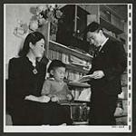 The Buddhist priest and store keeper for the Japanese evacuees at Picture Butte, Alberta, lives in a home well stocked with books. Here he is with his wife and youngest son. [1945/06/16-1945/06/28]