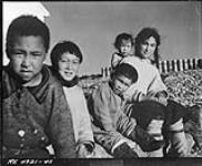 Five Inuit children seated outside [Left to right: unknown boy, Johnny (Kavavow?) Lyall, Pat Napatchee Lyall, Bella Ningyooga Wilcox (née Lyall) carrying baby Betty Novalinga Brewster (née Lyall), probably at Fort Ross, Nunavut] [Summer 1948].