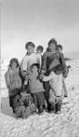 Group of Inuit women and children [Left to right, front row: Akittiq is crouching on the ground, Mukpa is standing in the centre and Oqadluk is on the right. Left to right, back row: Regilie, Killaq is carrying Josie Enuaraq and Paka is carrying her son Joanasie Simonie (woman on right, holding little the girl's hands)] October 1, 1943.