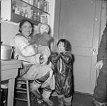 Woman holding a baby on her lap with a little boy standing next to them in the kitchen of the Hudson's Bay Company [The woman is Sarah AmakLak Haulli, from Hall Beach (Sanirajaq). The boy could either be Isaac Tullik Haulli or David Maniq Haulli and the little girl is Siqpaapik.] 1953.