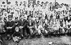 Pioneer Ukrainian settlers from Bukowina in front of St. Michael Ukrainian Greek Orthodox Church, Gardenton, Manitoba. Married women are wearing wrap-around head scarves; unmarried women are wearing floral cylindrical hats. 1915 1915.