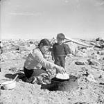 Inuit mother cooking bannock in frying pan over kerosene lamp fire August 1946.