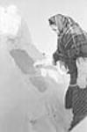 An Inuit woman wearing a plaid shawl standing beside a mound of snow at Inukjuak, Nunavik (Quebec) [she has been identified as Anna Matiusie, the mother of Annie Matiusie] 1947-1948.