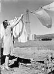 Mrs. Don Carr of Turner Valley is hanging out her weekly washing. An oil well derrick can be seen in the background June 1945
