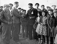 First Nation man wearing a uniform with a medal shaking hands with a non-First Nation man surrounded by a large group of First Nations men and children, unknown location, Northwest Territories. 1947.