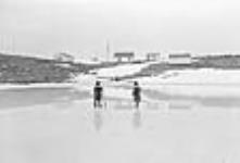 Inuit boy and girl standing on ice with buildings in the distance [between 1948 and 1953].