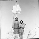 Inuit man and woman with pole where sealskins are hanging (out of the reach of the dogs) 1949