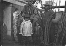 C.G.S. ARCTIC expedition. Kaktoo family on board [Qaktu (far right), his wife, Oolayoo (centre carrying an infant) and their family. Their daughter Oopikjuya (Rhoda) Allooloo is the girl on the far left. The man with the binoculars is probably William H. Grant. This picture was most likely taken en route to Devon Island.] 1922.