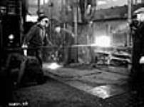 Un homme insère une pièce de métal moulée dans un grand four pour la chauffer avant qu'elle soit insérée dans des presses géantes. Le métal est chauffé à blanc Dec. 1939