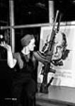 À l'usine de fusils-mitrailleurs Bren de la John Inglis Co., Veronica (Ronnie) Foster, employée de cette société et connue sous le nom de « la fille au fusil-mitrailleur », pose avec un de ces fusils terminé devant une affiche de Winston Churchill 10 May 1941