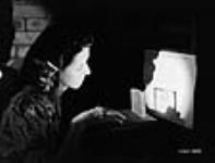 Afin de détecter des défauts, une ouvrière conduit un essai sur du verre au moyen de lumière transmise et d'un comparateur optique sur un écran à l'usine de la filiale d'instruments d'optique de la Canadian Arsenals Ltd July 1941