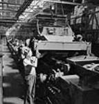 Des ouvriers guident la carrosserie d'épais blindage sur le châssis d'un véhicule de reconnaissance sur roues sur la chaîne de montage de l'usine de la compagnie Ford du Canada 2 July 1942