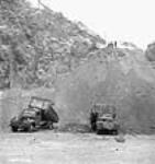 Trucks fill in the new road to the top of the tailrace excavation while bulldozers on top push earth down and workmen scatter the earth with shovels; Shipshaw Power Development project janv. 1943