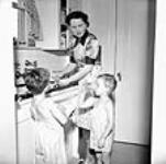 Mme Jack Wright lave la vaisselle pendant que Ralph Wright la sèche et David Wright boit un verre d'eau Sept. 1943