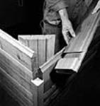 Un ouvrier tient une planche en bois de cèdre pour montrer l'assemblage àrainure et languette utilisé dans la construction de maisons préfabriquées mai 1944