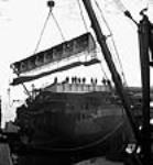 Lors de la construction de porte-avions d'escorte au chantier naval de Burrard, vue d'un mât de charge au bout duquel est suspendue la section arrondie du pont d'envol mai 1944