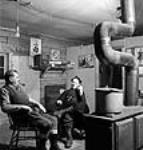 Provincial Government Sanitary Inspector, J.A. LePage of Montreal, Quebec and Father Edward Meilleur, O.M.I. smoking a pipe in front of a woodstove on a Sunday evening before service at the lumber camp cookhouse Mar. 1943