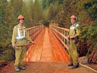 Two fire fighters standing in front of one of the Kettle Valley Railway trestles. The trestle is soaked in a red-coloured fire retardant. 14 of the 18 trestles in the Myra Canyon (just outside of Kelowna) were lost to the forest fire in August and September 2003 Aug 2003