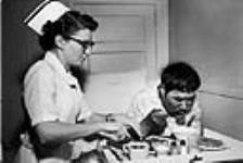 Miss E. On??s, Director of Nursing at hospital in Frobisher Bay, helps a patient eat his meal by cutting up his food 1959