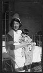 Nurse Madeline Stevenson sitting on a bed with an infant. Probably at the Carleton General Protestant Hospital ca. 1924.