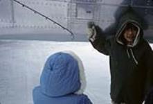 Inuit child standing beside a plane and talking to another Inuit child wearing a blue parka [graphic material] May 1965.