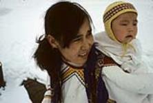Young mother and her baby: the mother is wearing an amauti and her baby is wearing a yellow hat [Damaris Ittukusuk Kadlutsiak was the first wife of Josiah Kadlutsiak of Igloolik. She is packing her son, Mike Kadlutsiak] [graphic material] May 1965.