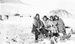 Four Inuk children sitting on a wooden structure in the snow with a large pile of logs and a ship in the background n.d.