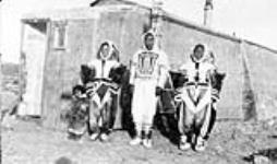 Two Inuit women, a child and a man wearing traditional parkas from the Kugluktuk region standing in front of a building [ca 1930]