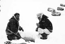 Two Inuit men ice fishing with sled dogs sleeping in the background n.d.