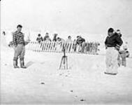 Two Inuit men standing in snow, with a group in background n.d.