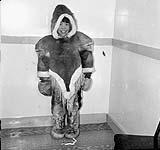 Inuk boy in a caribou parka, standing in a [nursing station] 1950
