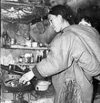 Inuk woman, carrying her baby in her amauti (parka), cooking bannock over primus stove in her home 1950