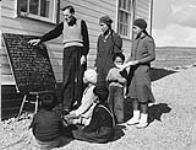 Anglican missionary, Reverend W. James. teaching his class of Inuit students how to read Inuktitut syllabics in Baker Lake (Qamanittuaq), Nunavut. [The woman in the polka-dot dress is Winnie Attungala, beside her is David Annanaut and Ruth Tulurialit; the girl with the white beret is Esa Tarraq.] 1948.