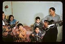 Two men playing guitars for a woman and three children at the Aklavik Mission Hospital, Aklavik, N.W.T [Women on bed is Ella Gordon, man on right is David Kalinek. Child in orange shirt is Peter Elanik Sr, and the boy in black and white is Wilson Malagana]] [ca. 1956]