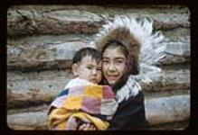 Young girl [Eunice Nasogaluak (Gordon)]holding an infant in front of a log building, Aklavik, N.W.T [ca. 1956]