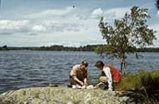 One man and one woman making preperations for a shore lunch on an island of the Lake of Woods, Ontario juillet 1950