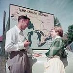 Deux voyageurs, un homme et une femme, consultent une carte routière devant un écriteau de l'autoroute transcanadienne à l'intersection des autoroutes 15 et 17 de l'Ontario, près d'Ottawa June 1952