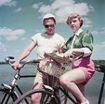 Deux cyclistes - un homme et une femme - consultent une carte routière sur le bord de la rivière Outaouais au parc Rockcliffe, en Ontario June 1952