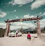 Man takes photograph of two women leaning against a car at the Great Divide of the Rockies at the summit of Kicking Horse Pass, Yoho National Park, British Columbia 1952