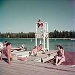 Two men and two women sit on a dock below a lifeguard at Clear Lake, Riding Mountain National Park, Manitoba  [Deux hommes et deux femmes assis sur un quai en-dessous d'un maître nageur au lac Clear, au parc national du Mont-Riding, Manitoba] 1952