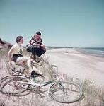 Man and woman seated on a beach with bicycle, Prince Edward Island National Park July 1953