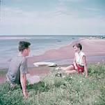Man and woman seated on the ground by the beach, Prince Edward Island National Park  [Un homme et une femme sont assis sur le sol près d'une plage, parc national de l'Île-du-Prince-Édouard.] juillet 1953