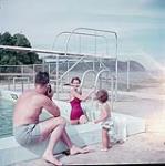 Man photographing a woman in red bathing suit climbing out of pool, small child watches, Fundy National park, New Brunswick juillet 1953