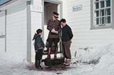 Un homme inuit et un jeune garçon inuit parlent à un agent de la GRC sur les marches d'un bureau de poste en hiver, dans les Territoires du Nord-Ouest [Nunavut] 1957