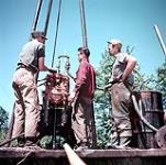 Trois hommes, tous membres d'une équipe de forage d'Hydro, se tenant près d'une grosse foreuse. Projet de documentation de la Voie maritime du Saint-Laurent. Cauhnawaga, Montréal 1954
