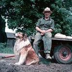 Male farmer sitting on a flat-bed trailer. Dog sitting in the foreground. St. Lawrence Seaway Documentation Project. Iroquois 1954