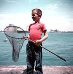 Jeune garçon portant une chemise rouge et tenant un filet de pêche. Projet de documentation de la Voie maritime du Saint-Laurent. Port Colborne 1954