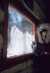 Woman (possibly Rosemary Gilliat Eaton) wearing a winter coat with a fur-trimmed hood and using photographic equipment to make images of frost on the windows. Shilly Shally Lodge, Gatineau Park 1963