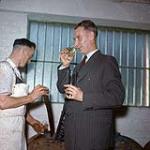 Homme vêtu d'un complet goûtant du whisky pendant qu'un autre homme portant un tablier décante le whisky d'un baril de bois [between 1955-1963]