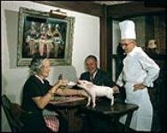 Colonel and Mrs. G.E. Leprohon, patrons of Au Lutin, Montreal, feeding the restaurant pig with baby's bottle. Owner B.J. McAbbie is shown at right. Le colonel et Mme G.E. Leprohon, clients du Au Lutin, Montreal, nourrissant le cochon du restaurant avec un biberon. Le propriétaire B.J. McAbbie est vu à la gauche. 1949.
